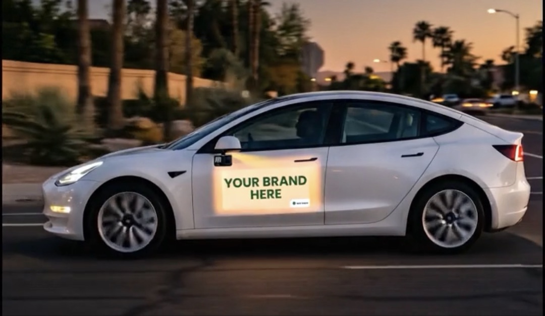 White Tesla Model 3 driving through Phoenix at golden hour with a projected 'YOUR BRAND HERE' ad on its side door panel.